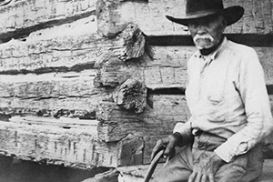 A man with a white mustache and a cowboy hat sits outside a log cabin